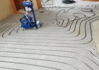 Zdjęcie przedstawia proces instalacji ogrzewania podłogowego. Na szarym podłożu wyfrezowano rowki, tworzące pętle. Widać także niebieską maszynę MV230, służącą do frezowania rowków w posadzce pod te rurki.