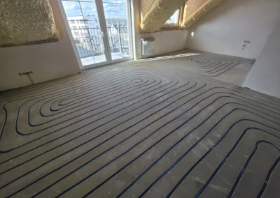 Węże ogrzewania podłogowego ułożone w spiralnym lub meandrowym wzorze na surowej betonowej posadzce w jasnym pomieszczeniu z drzwiami balkonowymi lub dużymi oknami.